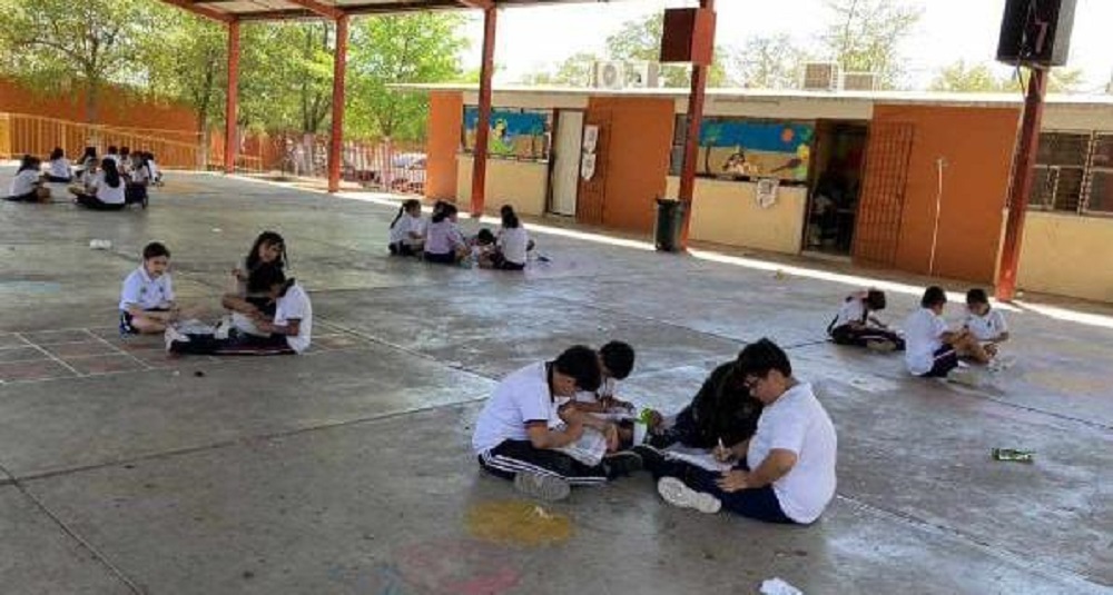 (FOTO) En el piso, bajo la techumbre, toman clases niños de la escuela